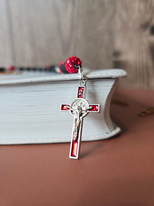 A black rubber coated glass beads and red clay beads with red glass rhinestones laying down on a white bible with a sliver crucifix with red enamel and St. Benedict medal behind Jesus as the mine focal point.