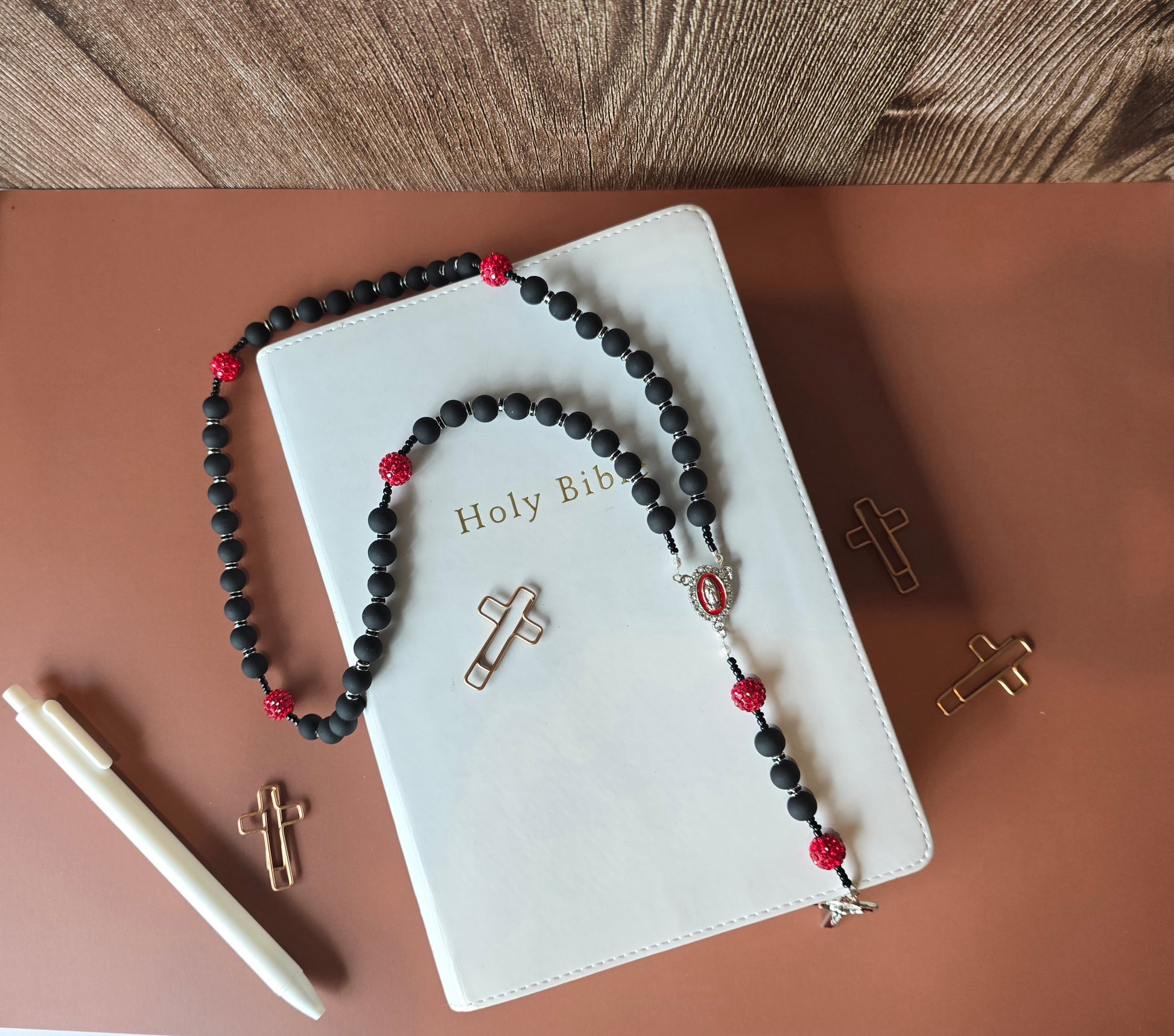 A black rubber coated glass beads and red clay beads with red glass rhinestones laying down on a white bible with a sliver crucifix with red enamel and St. Benedict medal behind Jesus and the Our Lady of Guadalupe medallion with white rhinestones around her.