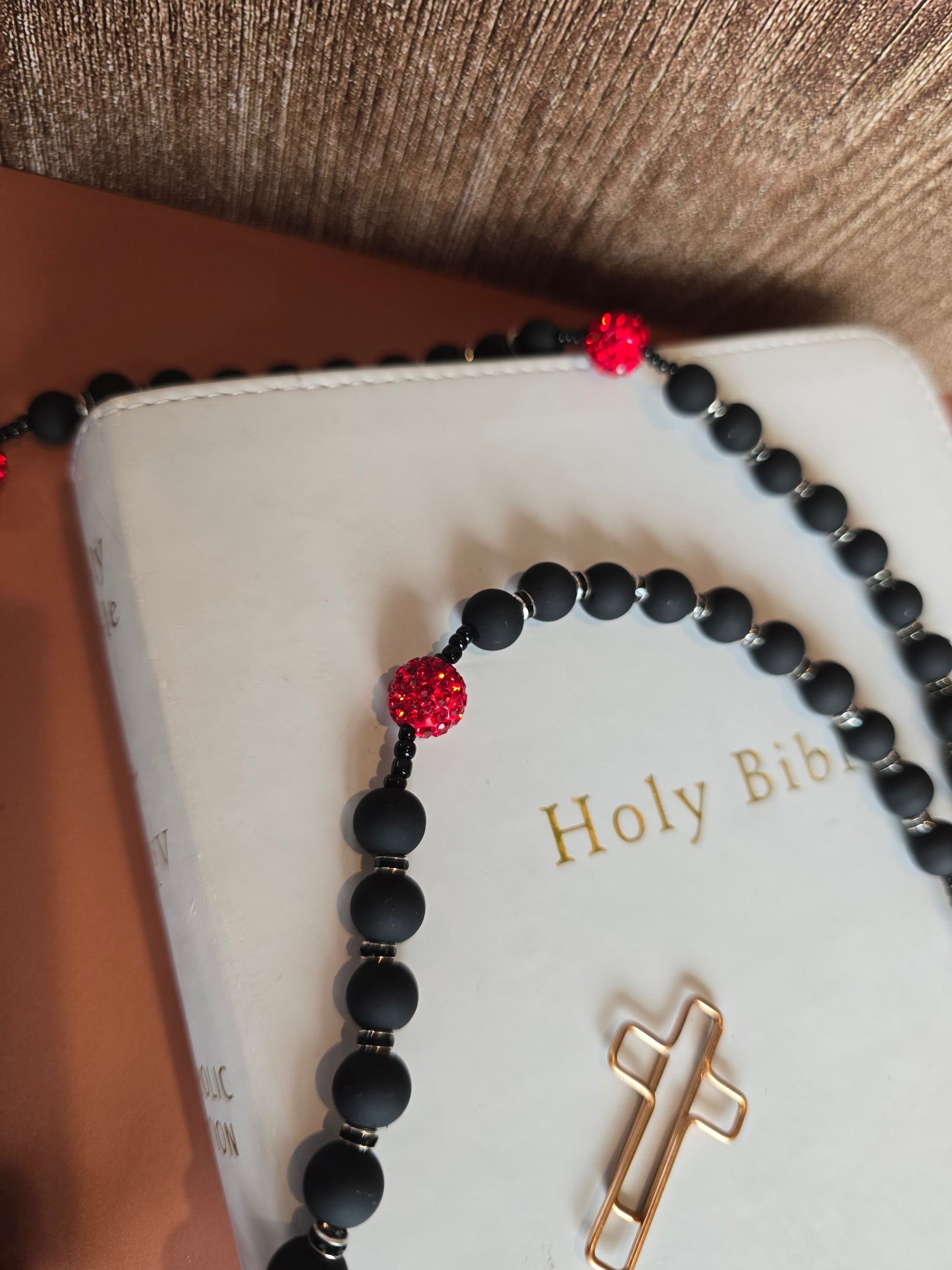A black rubber coated glass beads (Hail Mary) and red clay beads with red glass rhinestones (Our Father) laying down on a white bible with a focul point of our black rubber coated glass bead to show their matted texture as well as our red clay based beads with red glass rhinestones as we show case our black spacer beads.