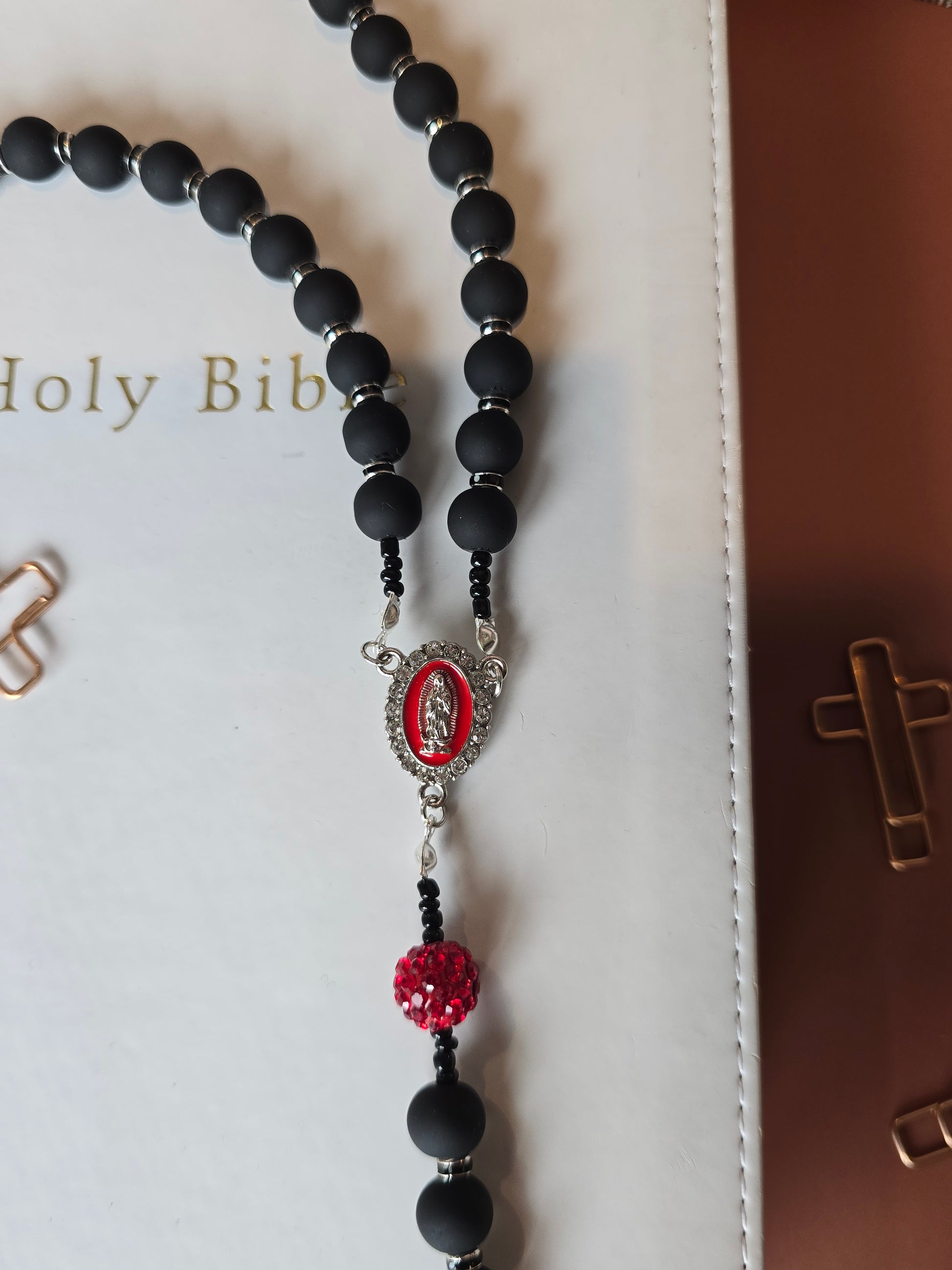 A black rubber coated glass beads and red clay beads with red glass rhinestones laying down on a white bible with a focul point of Our Lady of Guadalupe medallion with white rhinestones around her and our silver metal connectors.