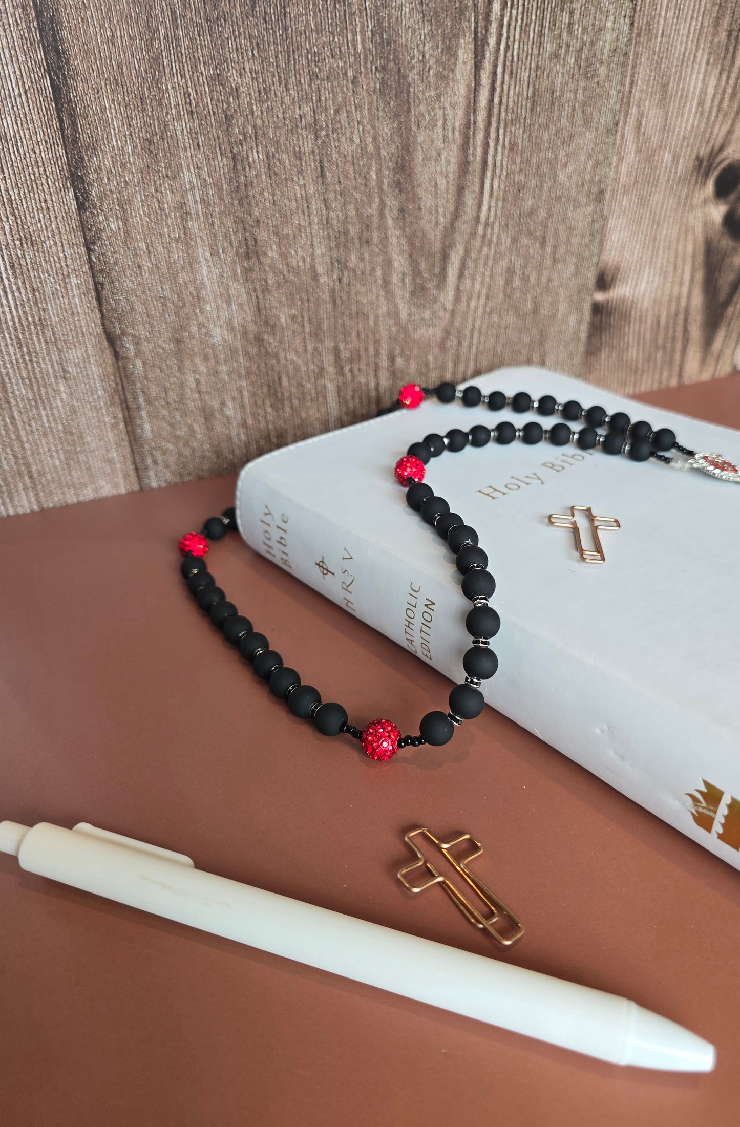 A black rubber coated glass beads and red clay beads with red glass rhinestones laying down on a white bible with a focul point of our black rubber coated glass bead to show their matted texture.